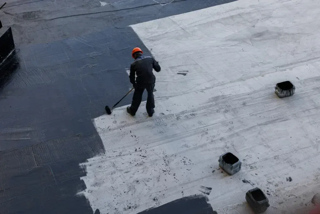 Person in orange hard hat painting a flat roof with a roller; dark gray and white paint.