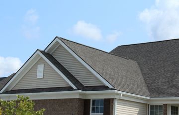 House with a brown shingled roof, beige siding, and a blue sky background.