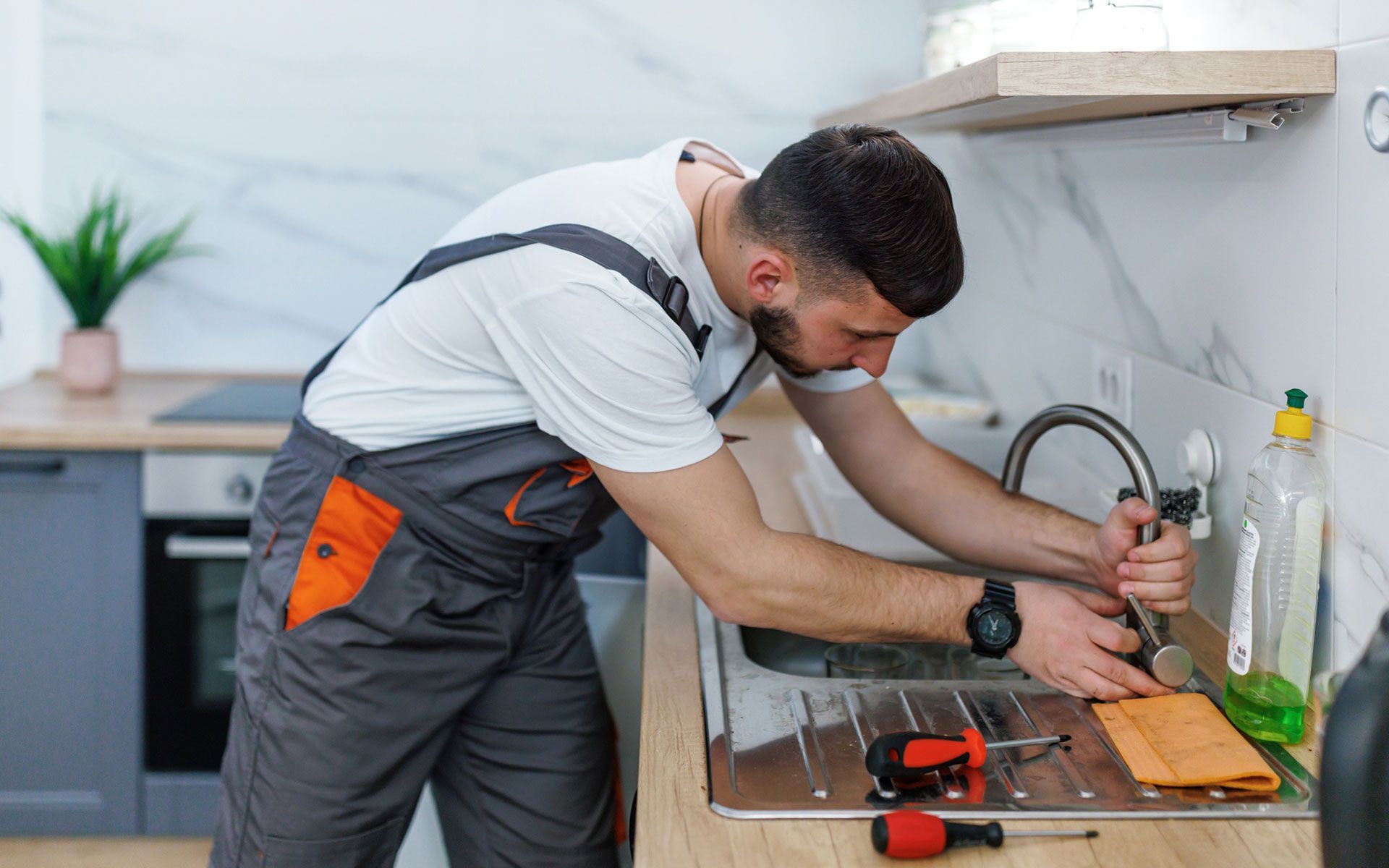 Plumber in gray overalls fixes a kitchen sink with a wrench. Tools and cleaning supplies visible.