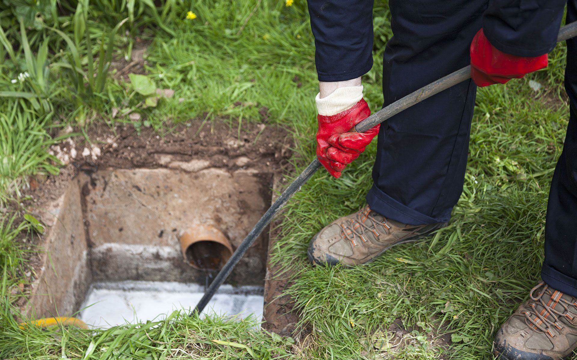 Person with red gloves using a rod to clear a drain in a grassy area.