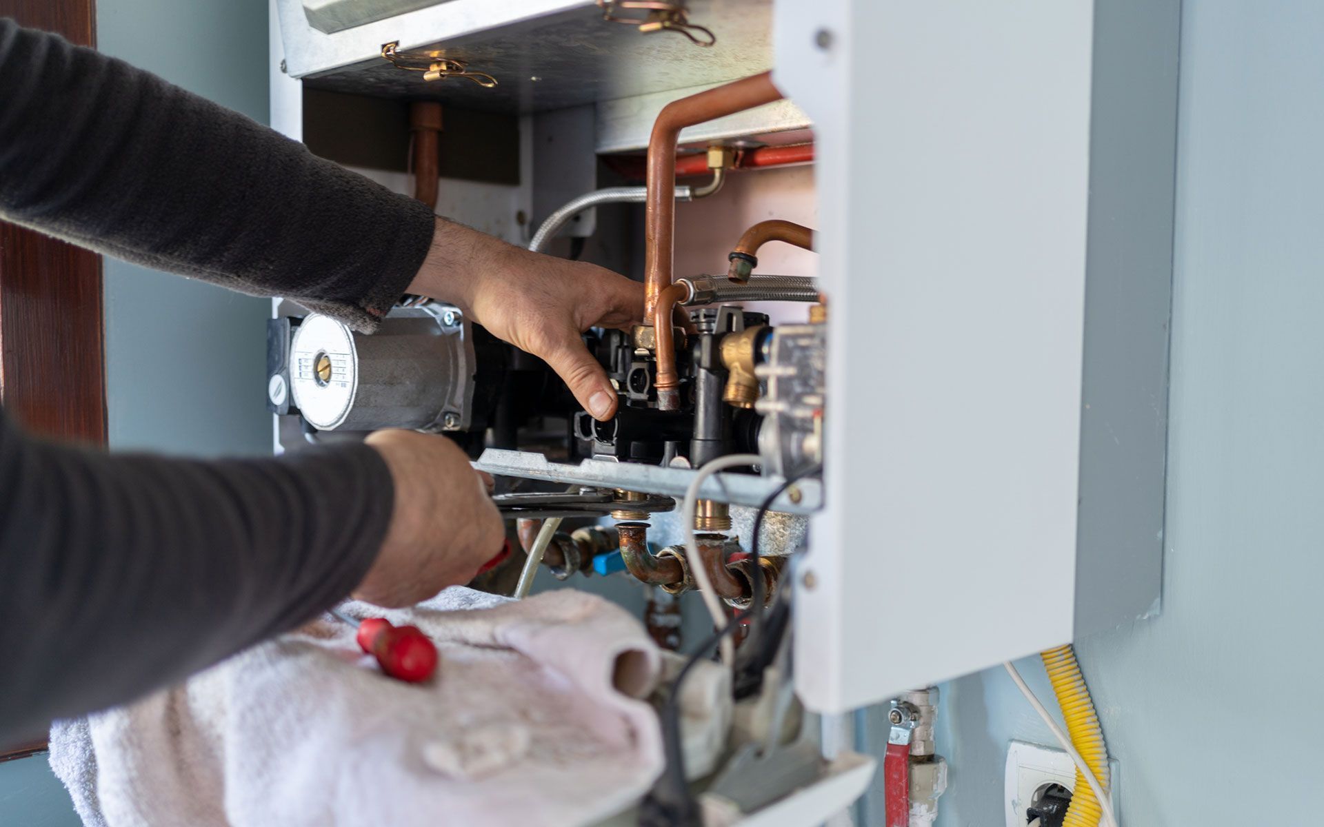 Person repairing a white gas furnace, using a screwdriver, indoors.