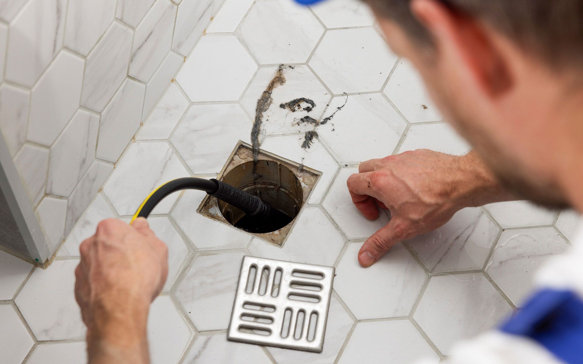 Person unclogging shower drain with a hose, dark gunk spraying on white hexagonal tiles.