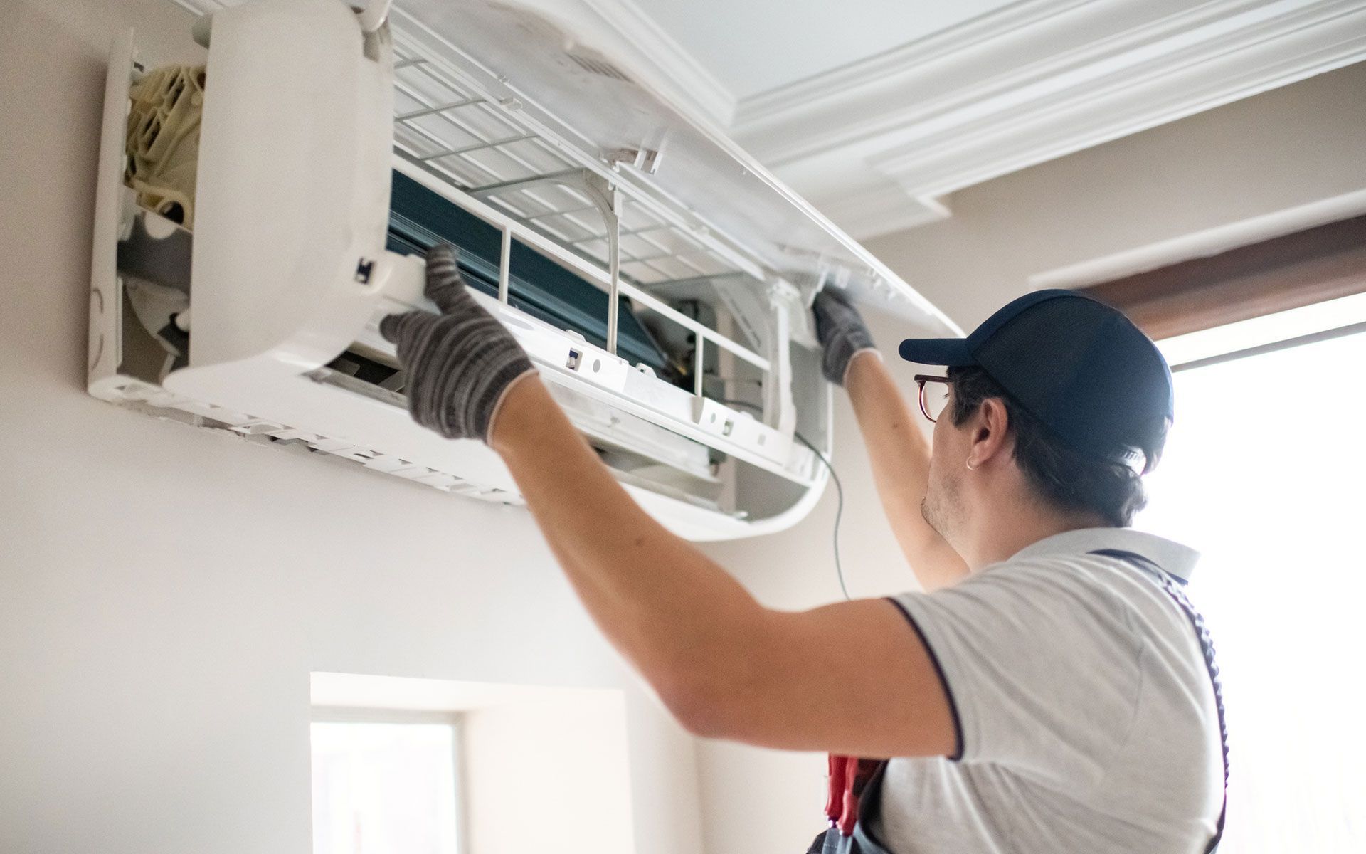 Person in cap and gloves servicing an air conditioner on a wall.