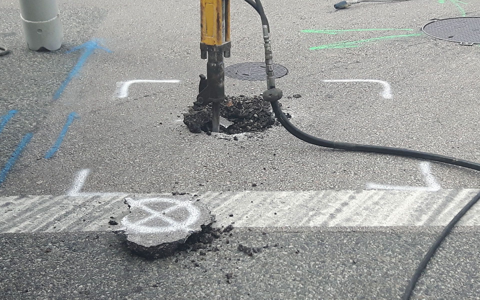 Asphalt being jackhammered, exposing concrete and causing debris. White and blue paint markings visible.