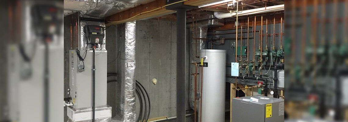 Plumber in gray overalls using a wrench to connect a flexible hose to a white water heater.