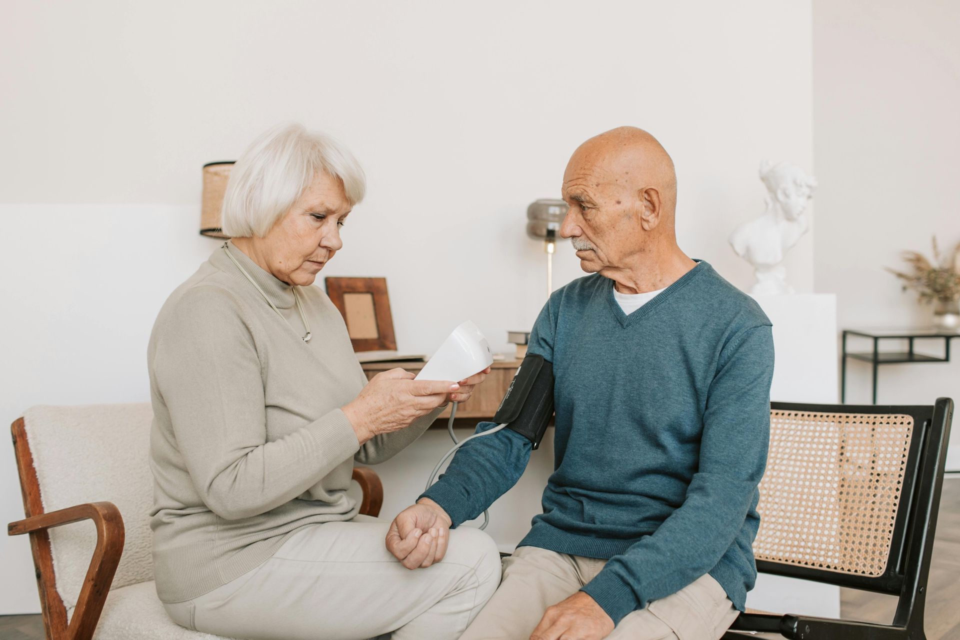 An elderly woman checks an older man's blood pressure using a monitor in their home.