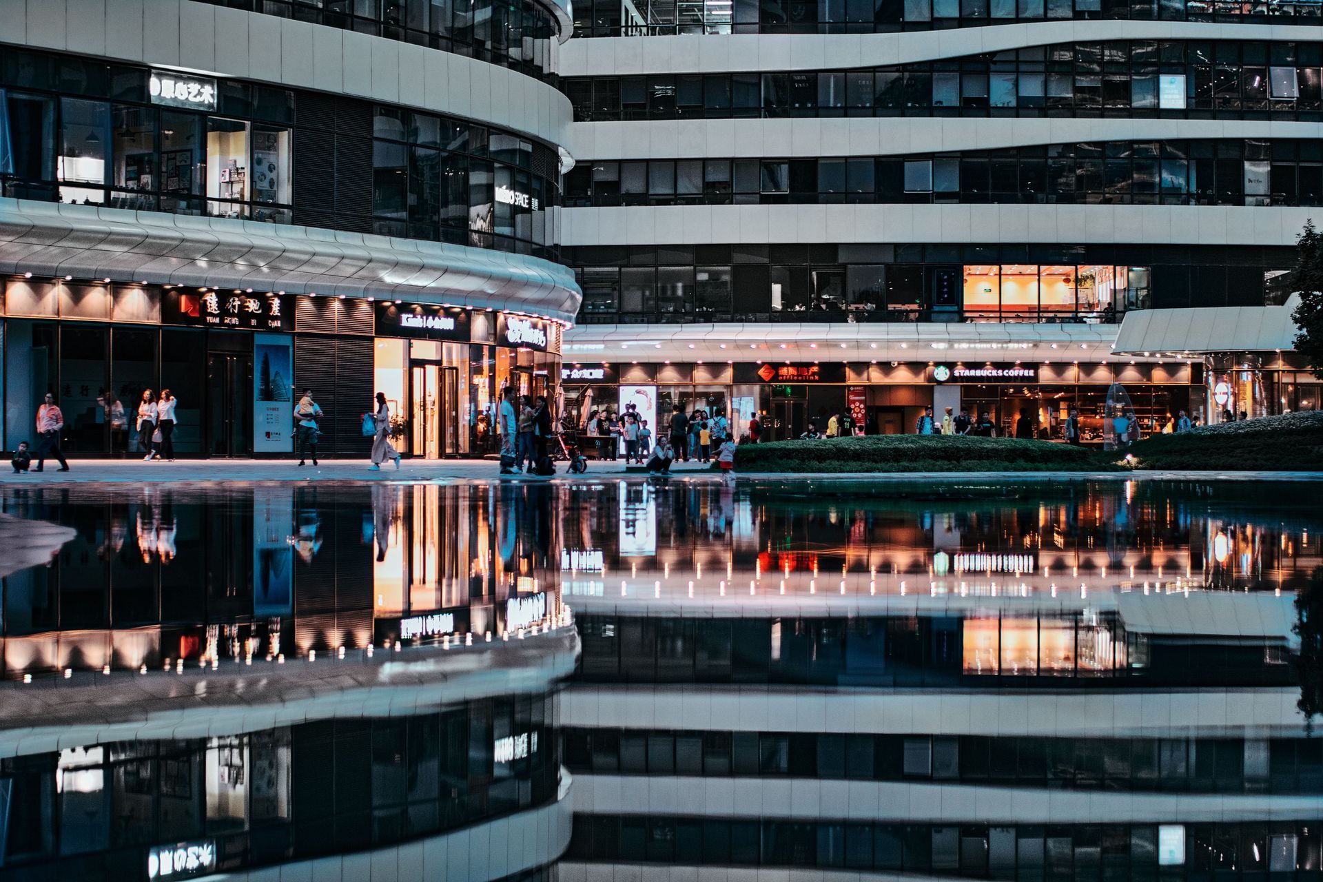 Office buildings with stores at the bottom floor and their reflection off of a man-made pond. There are people walking in front of the stores.