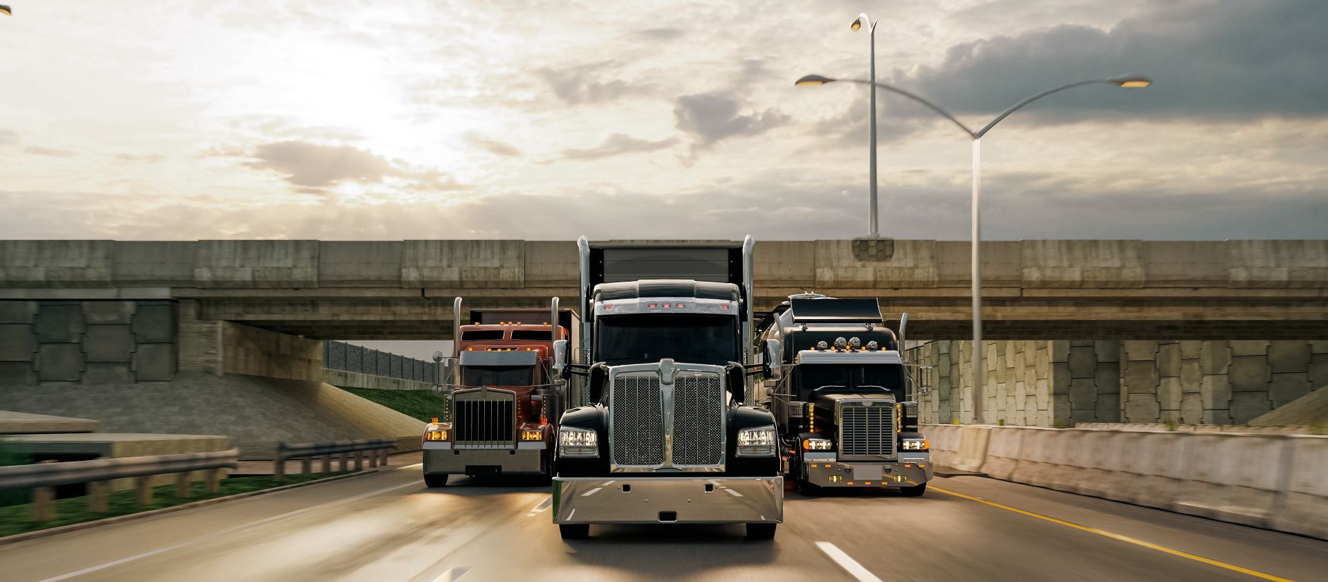 Three trucks driving toward the camera on a highway under an overpass at dusk