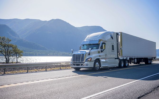 White semi-truck driving along a lakeside road with mountains in the background
