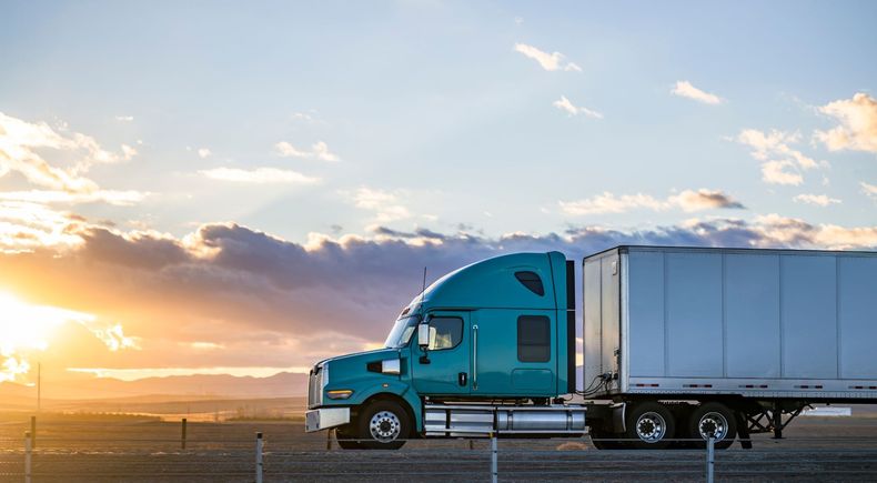 Blue semi-truck and trailer parked by a road at sunset with clouds and open sky