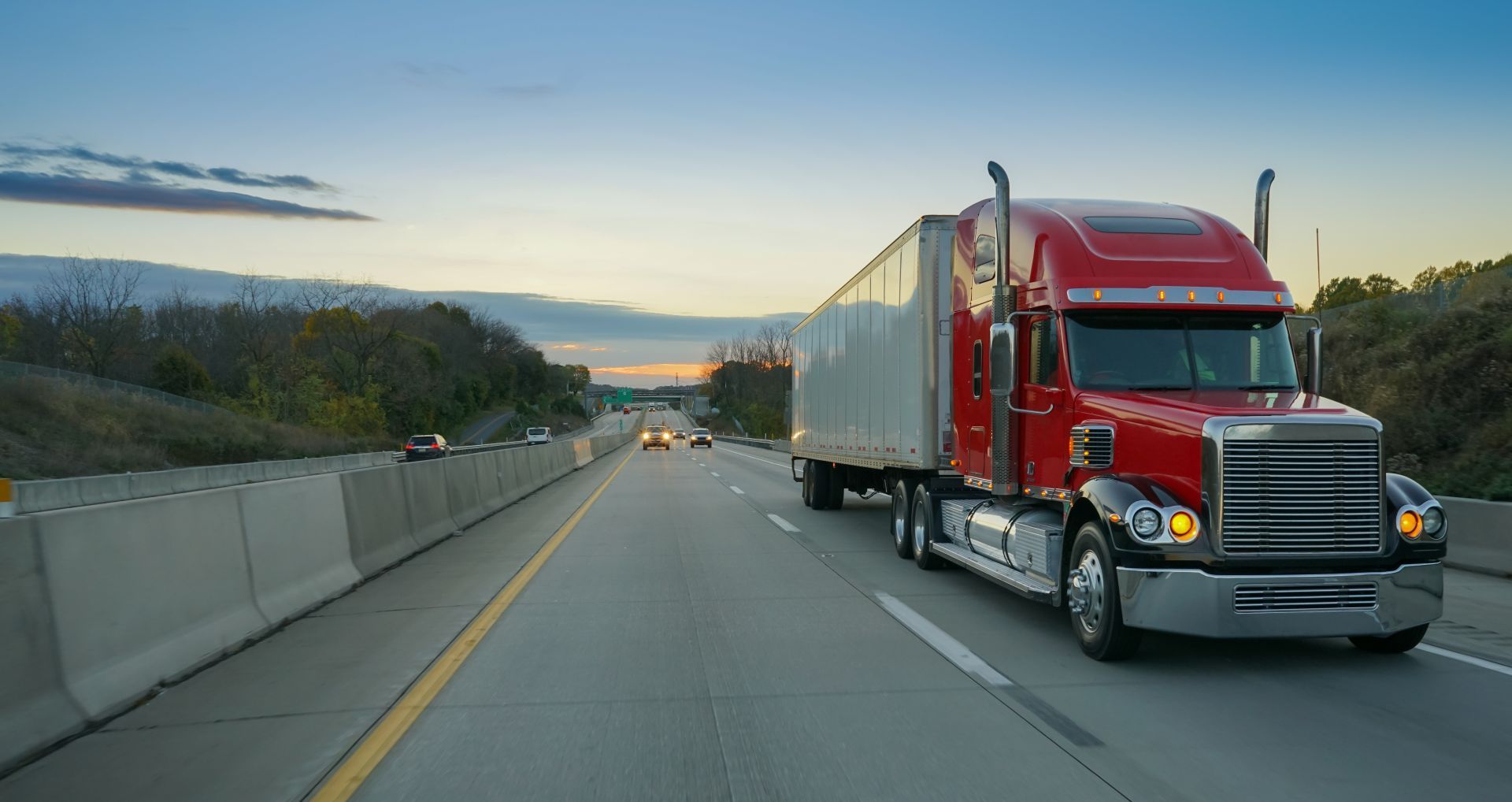 Red semi-truck driving on a highway beside a concrete barrier at dusk