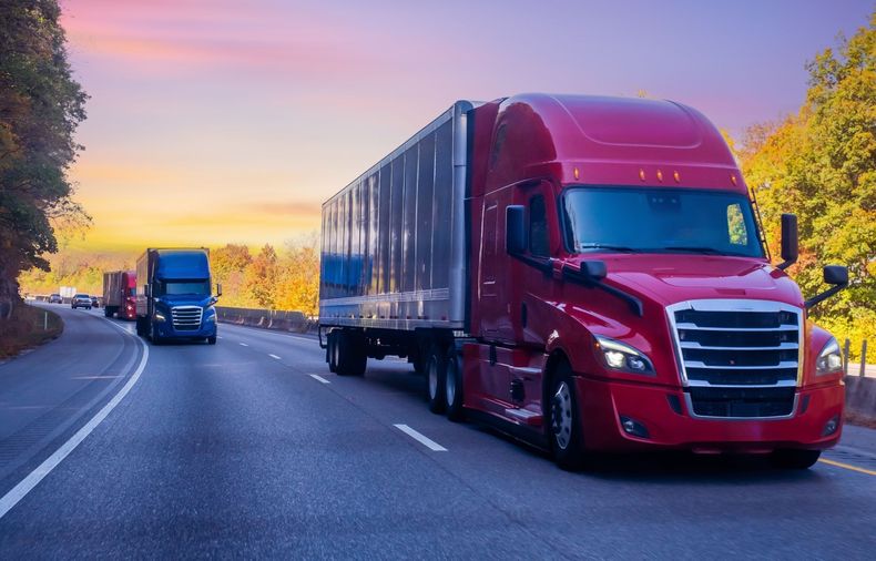 Red semi-truck driving on a highway at sunset, with another truck ahead and autumn trees beside the road