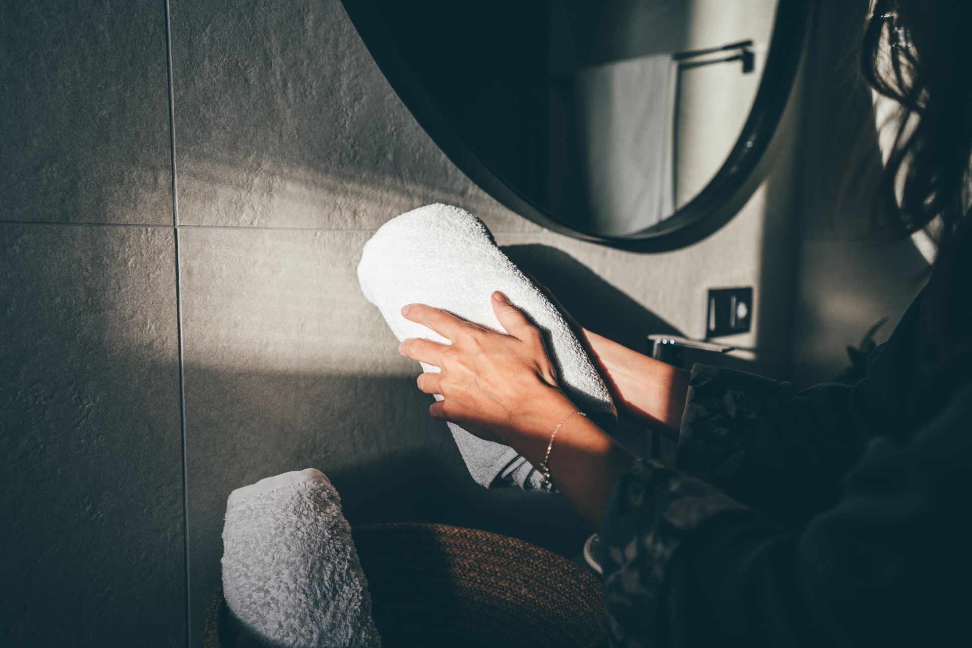 Persona che tiene in mano un asciugamano bianco in un bagno con uno specchio rotondo e luce solare.