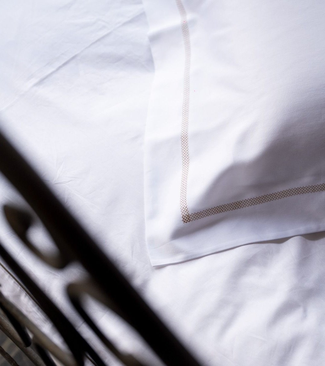 Lenzuola bianche con un cuscino bordato di marrone chiaro, viste parzialmente con una struttura del letto in metallo nero.