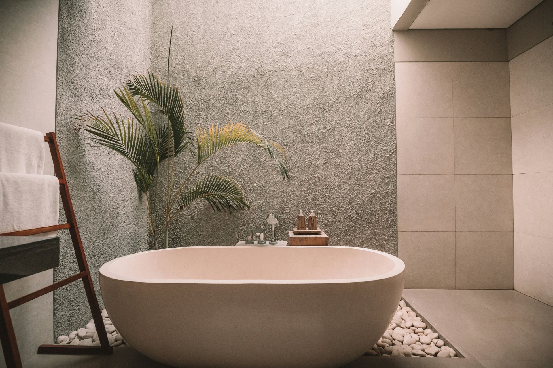 A single standalone tub on a small pebble flooring with beige tile work, a large plant, and a stone backsplash.