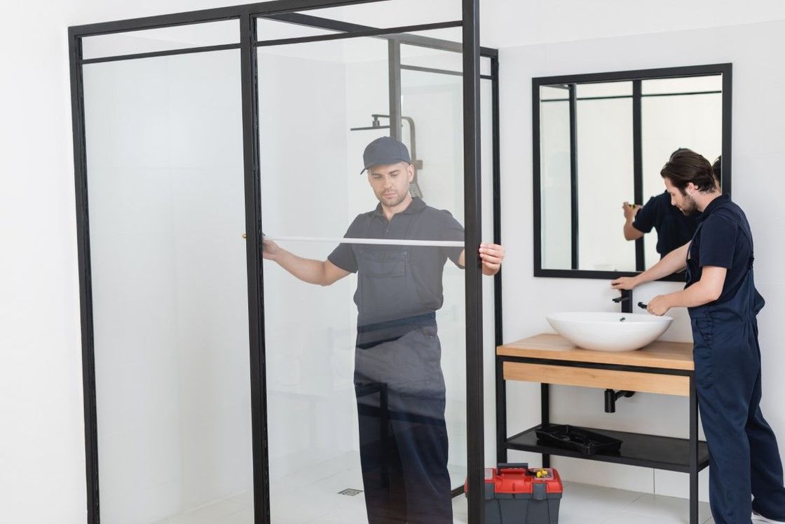 Two male workers in dark blue uniforms are assembling a modern-looking shower and bathroom sink.