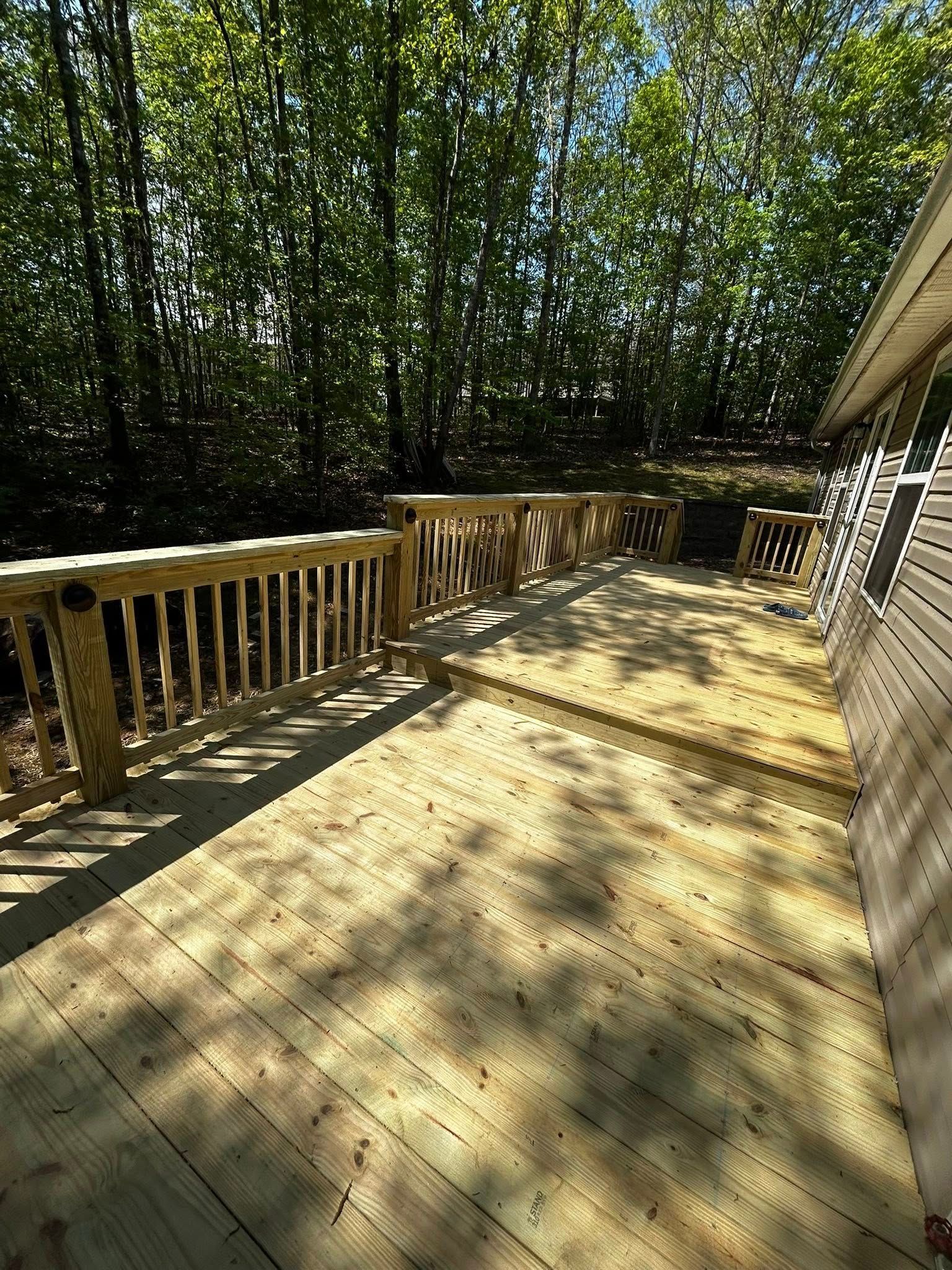 Wooden deck with railing, stairs, and attached to a house; surrounded by trees in the background.