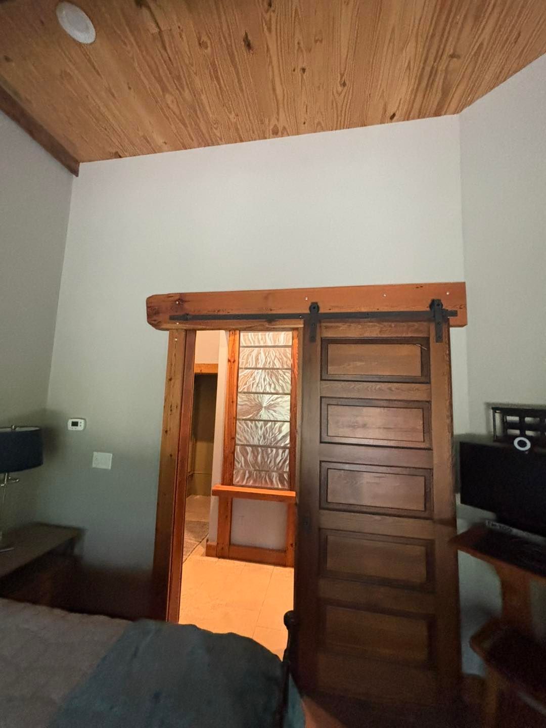 Wooden sliding barn door, leading to a room with a window, white walls, and a wood-paneled ceiling.