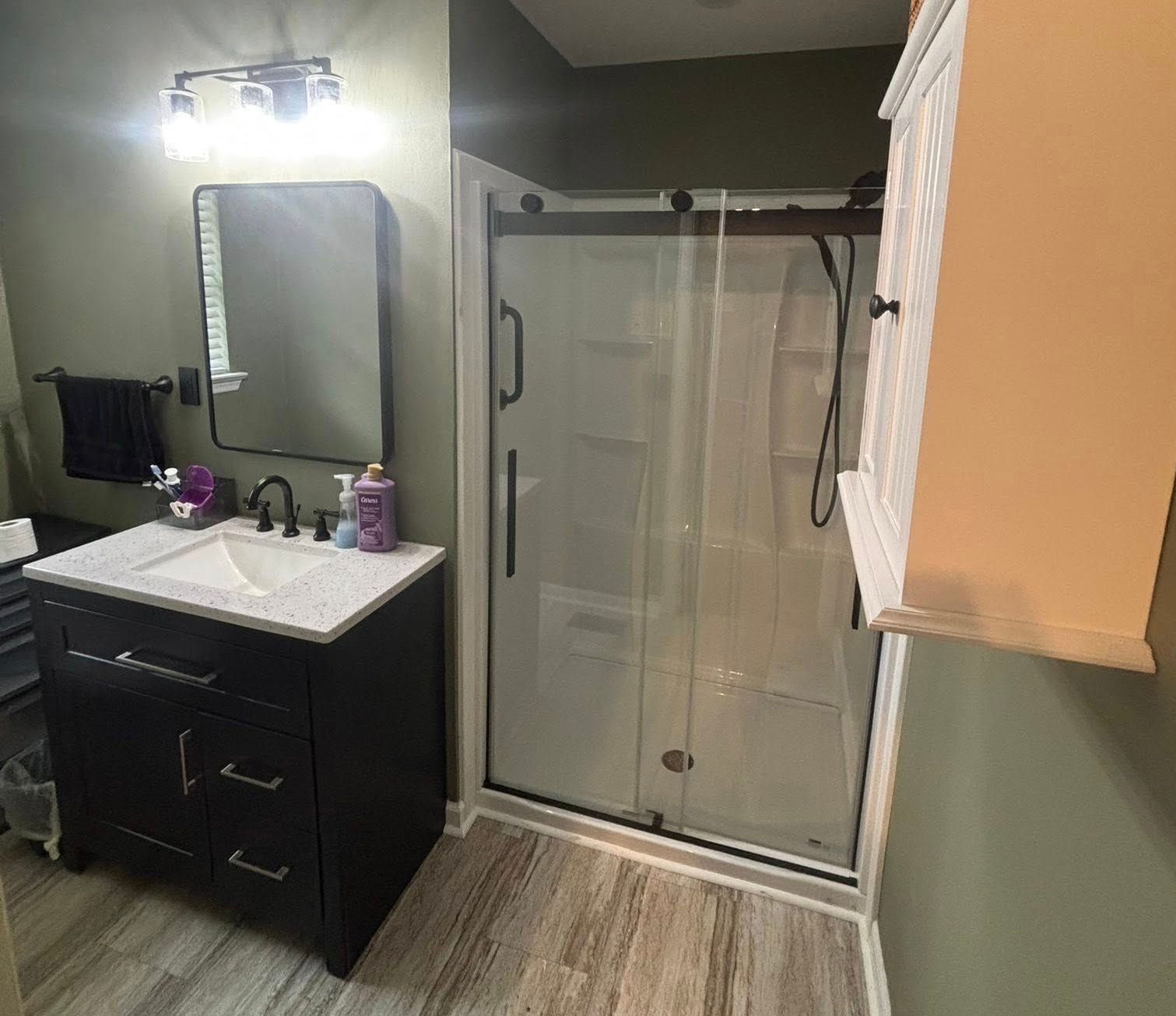 A modern bathroom featuring a dark vanity, a rectangular mirror, a glass-enclosed shower, and wood-look flooring.