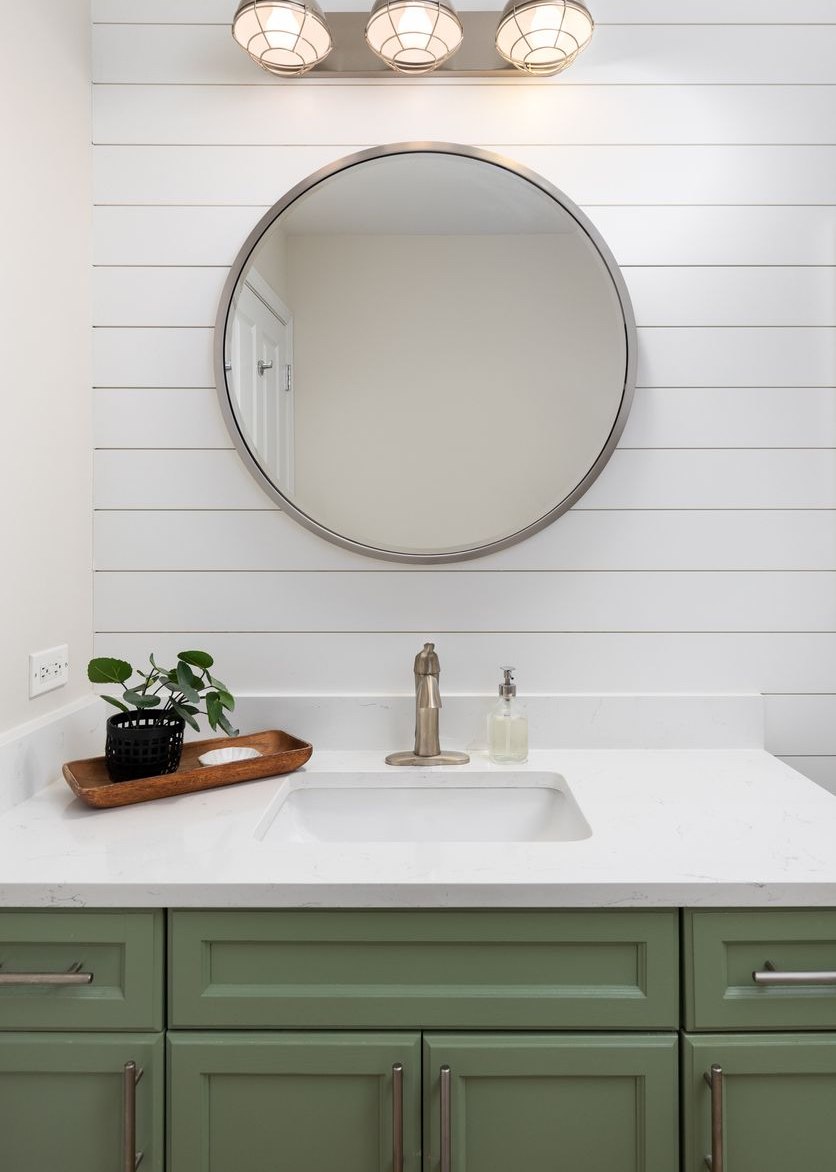 Green bathroom vanity with white countertop, round mirror, and shiplap wall.