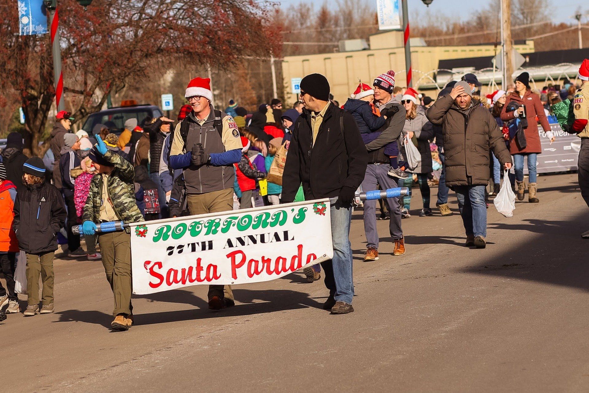 Roenford's 75th Annual Santa Parade with people wearing Santa hats, walking down a street in daylight.