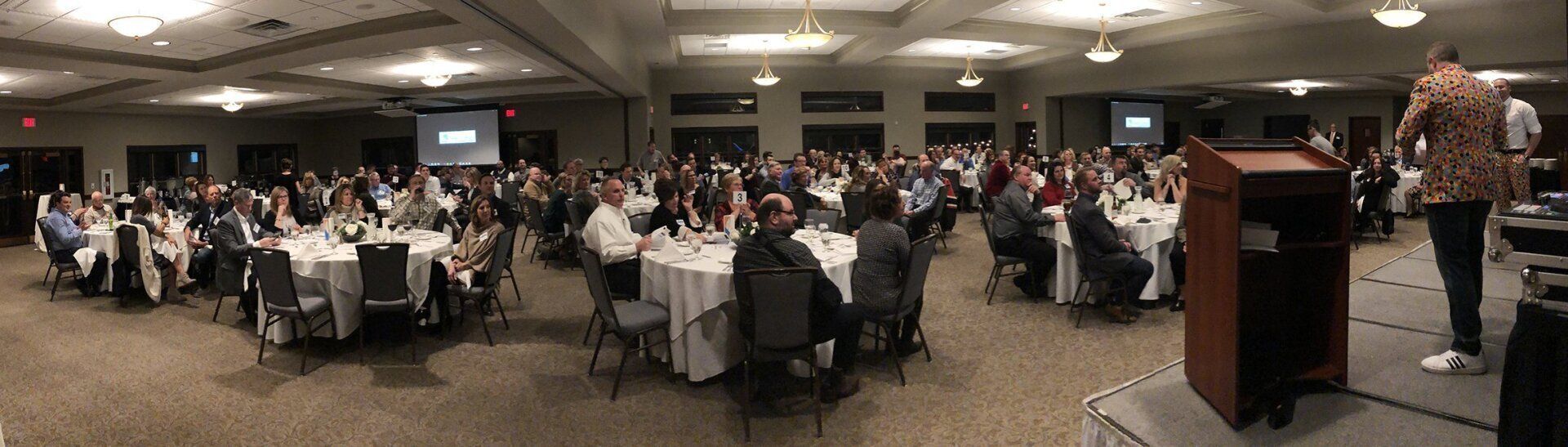 Large conference room with many people at tables, presenter at podium.