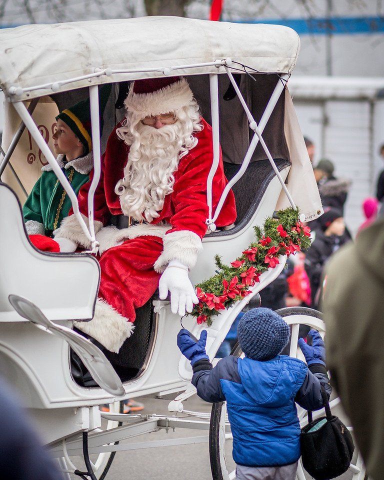 Santa in a white carriage waves to a child in a blue coat, garland on carriage.