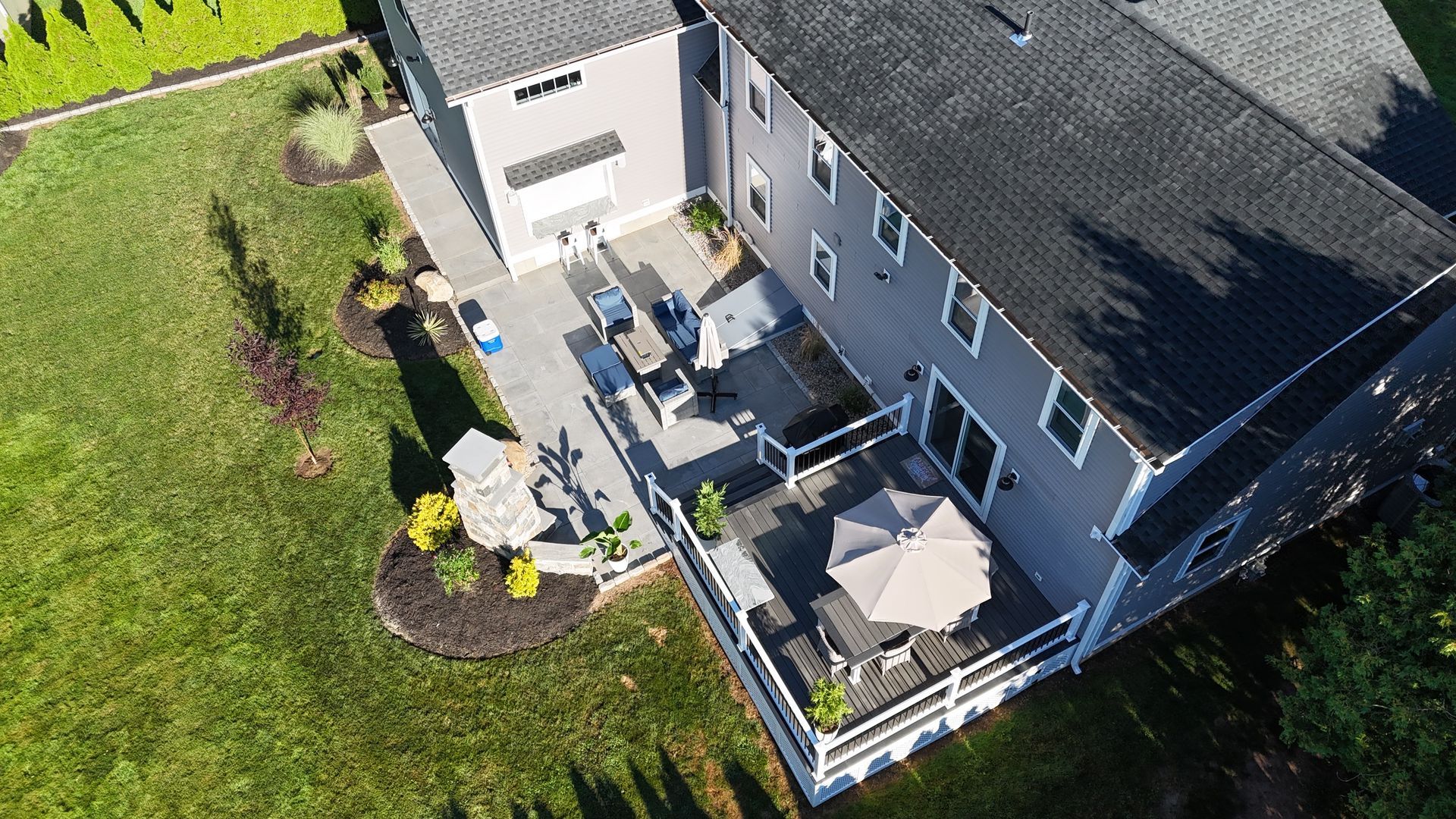 Aerial view of a gray house with a deck and patio, surrounded by green grass and landscaping.