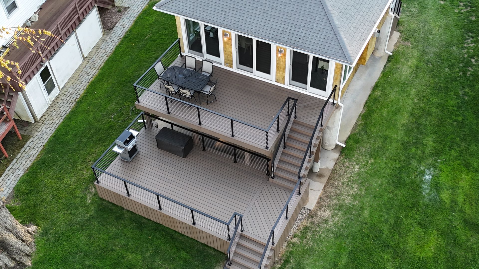 Two-tiered deck with seating, grill, and stairs attached to a yellow house with a gray roof, set on green grass.