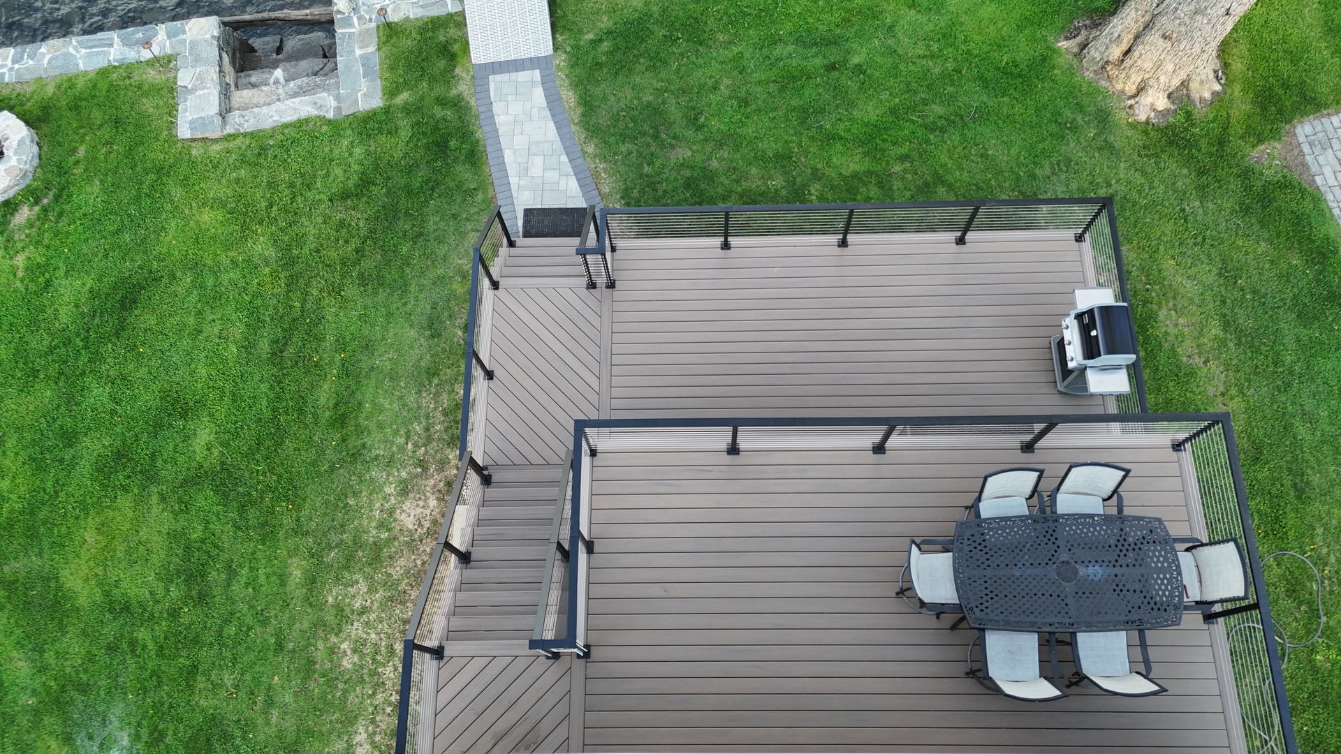 Overhead view of a composite deck with black railings, a grill, and a dining table with chairs, surrounded by grass.