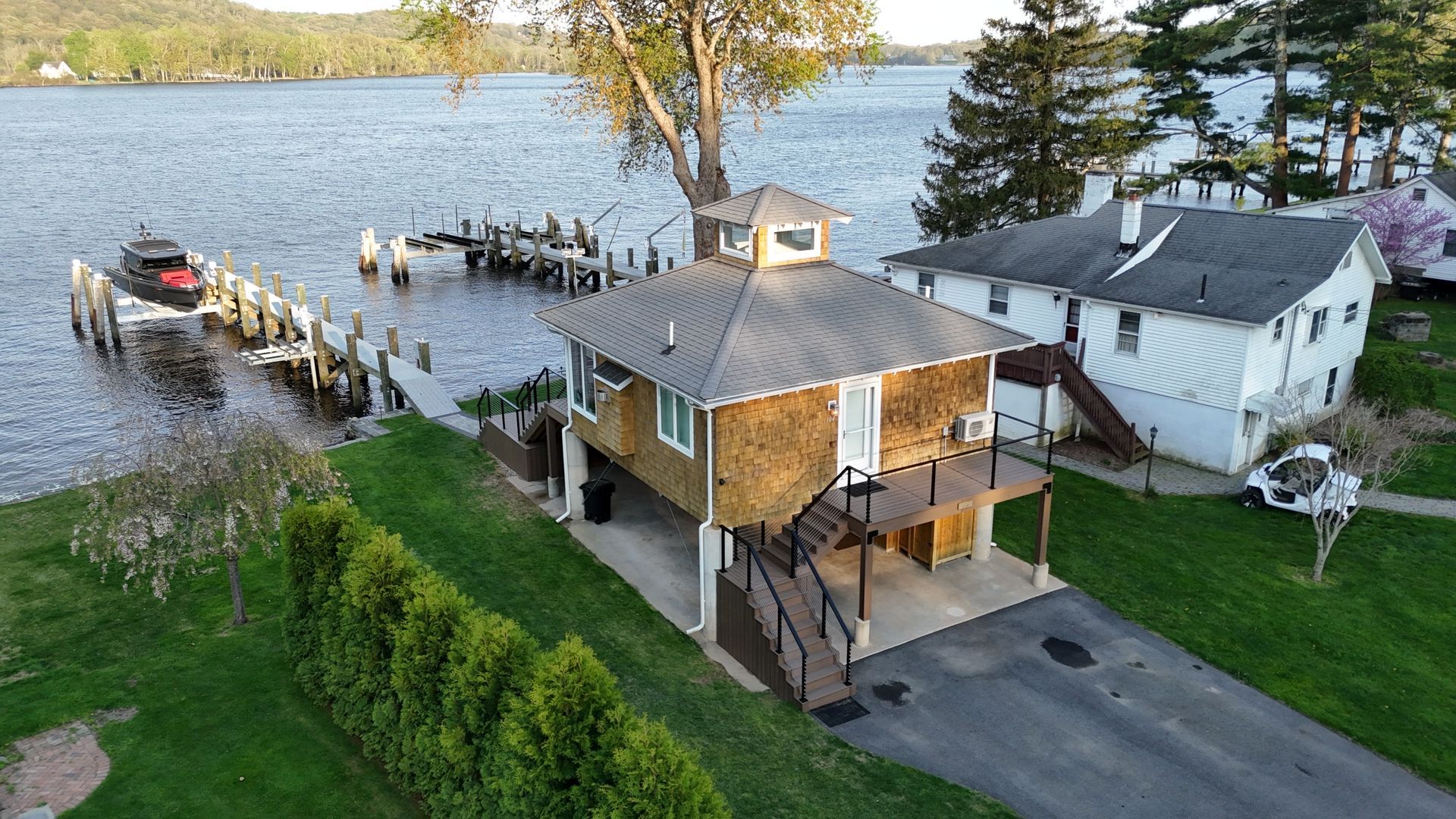 House with dock on water, brown siding, a small boat, green bushes, and two other houses.