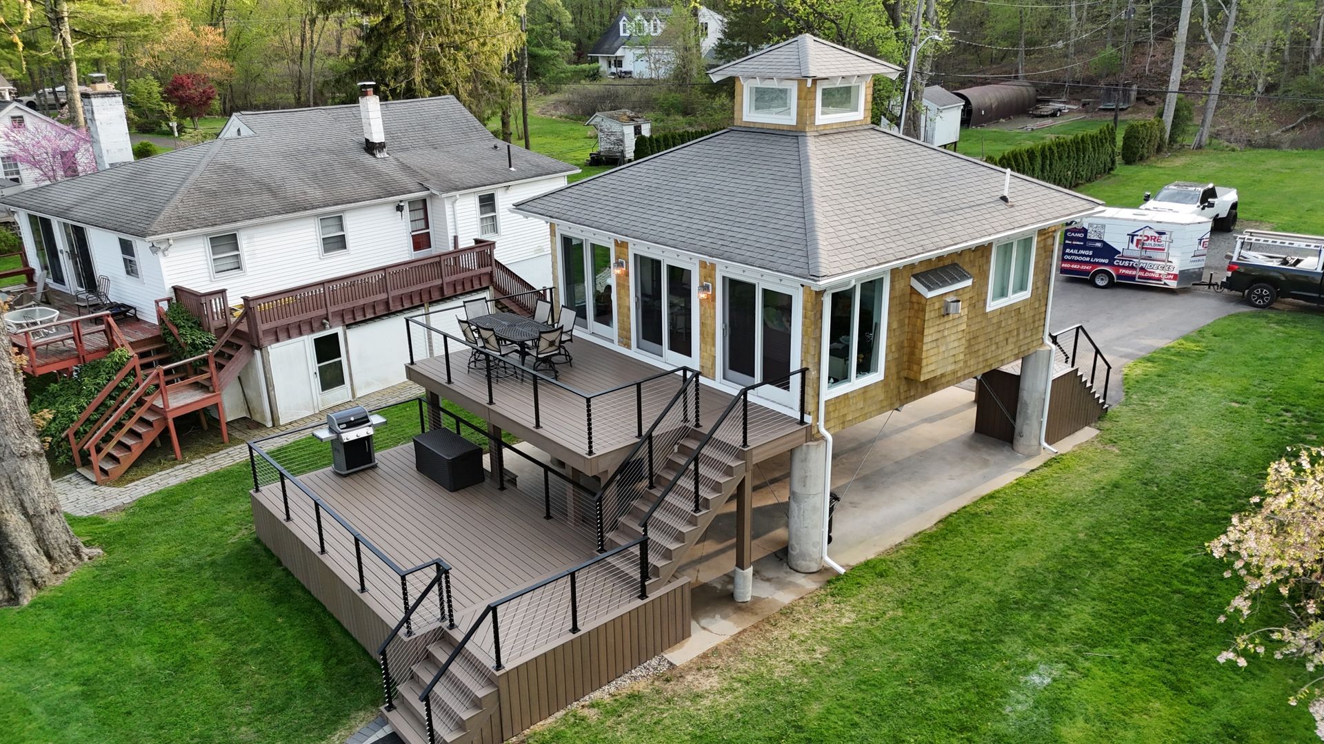 Two-story house with deck, and a smaller building, viewed from above, on green grass.