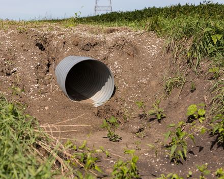 A corrugated metal drainage pipe sits at the base of a dirt embankment in a grassy field.