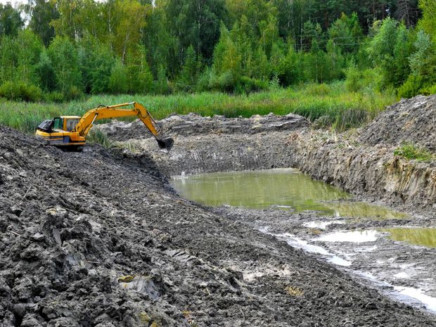 A yellow excavator works on a muddy, rocky embankment overlooking a small, murky pond near a forest.
