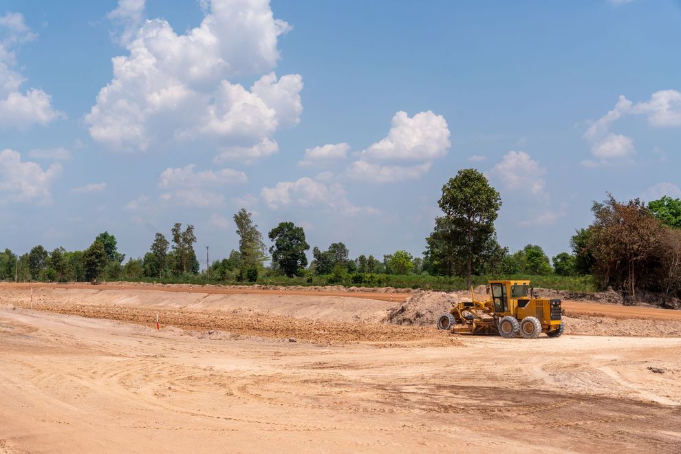 A yellow motor grader levels dirt on a large, sunlit construction site under a partly cloudy blue sky.