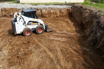 A white skid-steer loader works in a muddy, excavated dirt construction site.