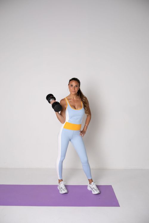 A woman is standing on a yoga mat holding a dumbbell.