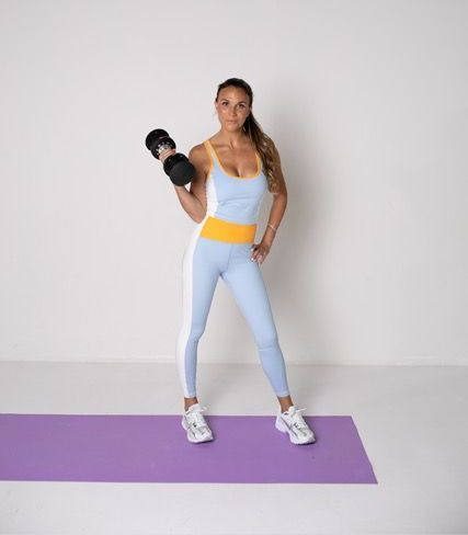 A woman is standing on a yoga mat holding a dumbbell.