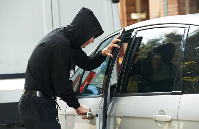 A person in a black hoodie is trying to break into a silver car.