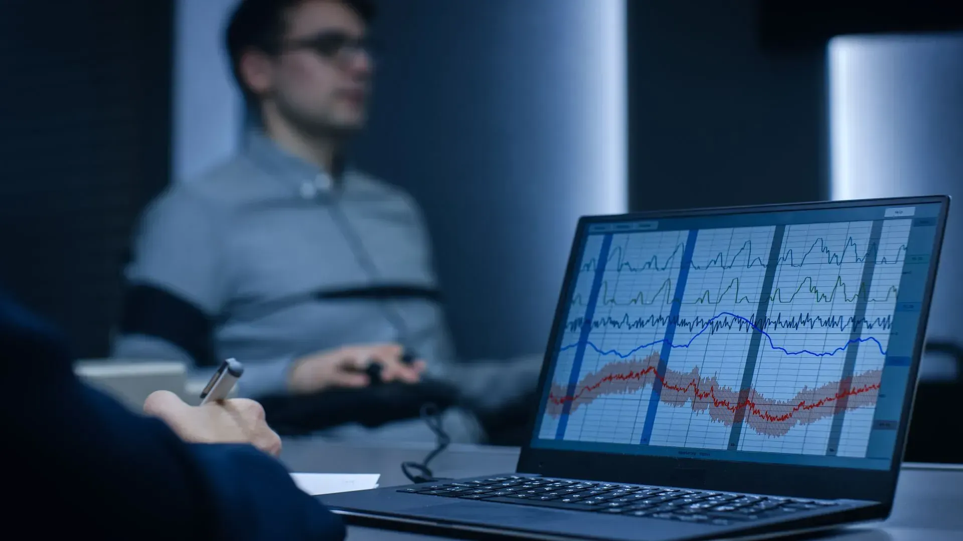 Laptop displaying graphs; person in gray shirt hooked up to sensors; interrogation room setting.