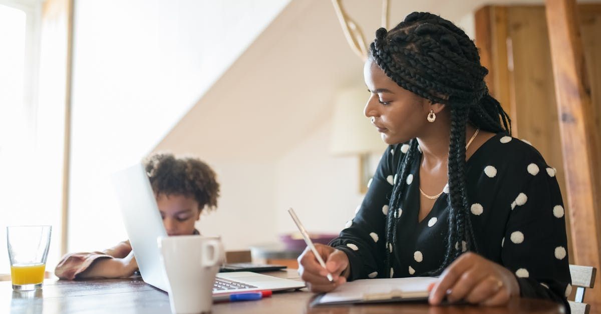 A woman is sitting at a table using a laptop computer while a child looks on.