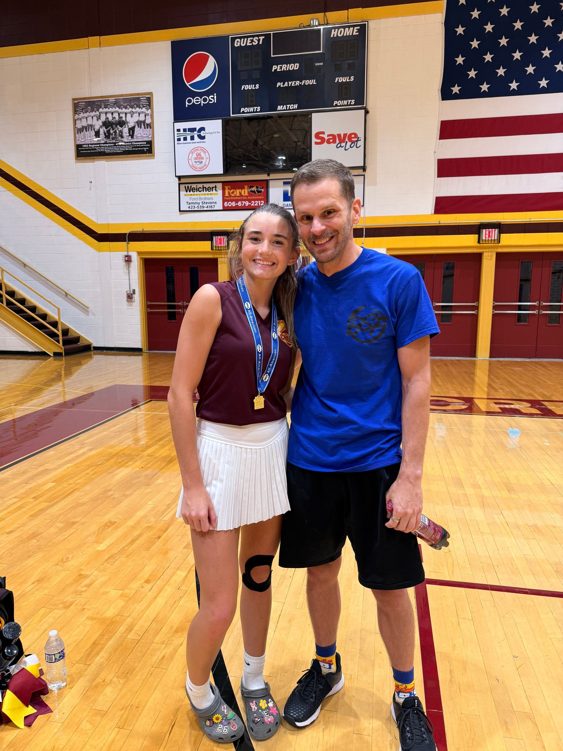 A young woman with a medal and a man pose in a school gym. She wears a maroon shirt, tennis skirt, and knee brace. He wears a blue shirt and shorts.