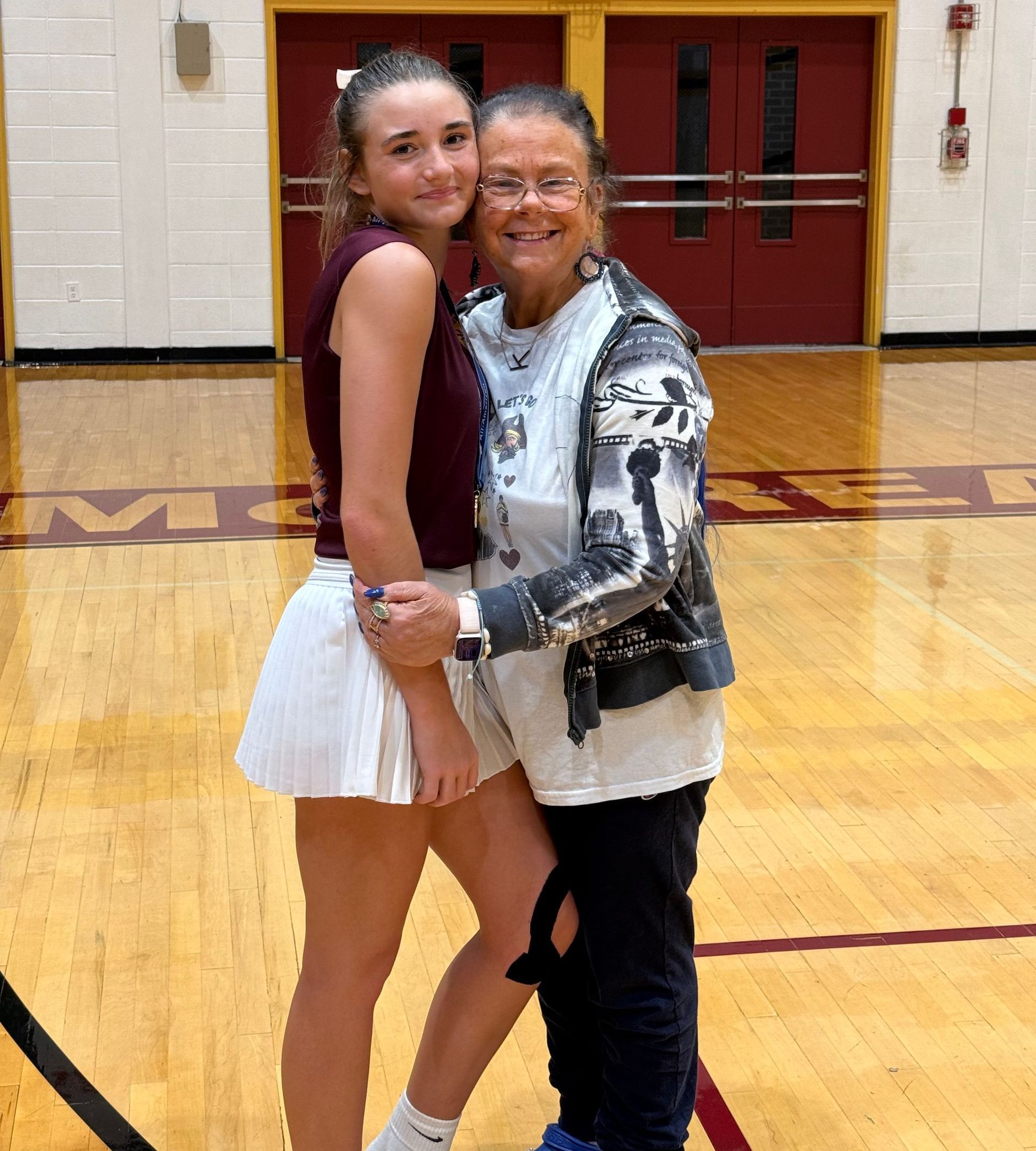 A young woman and older woman embrace on a basketball court. The younger woman wears a burgundy tank and white skirt, the older wears a tie-dye shirt.