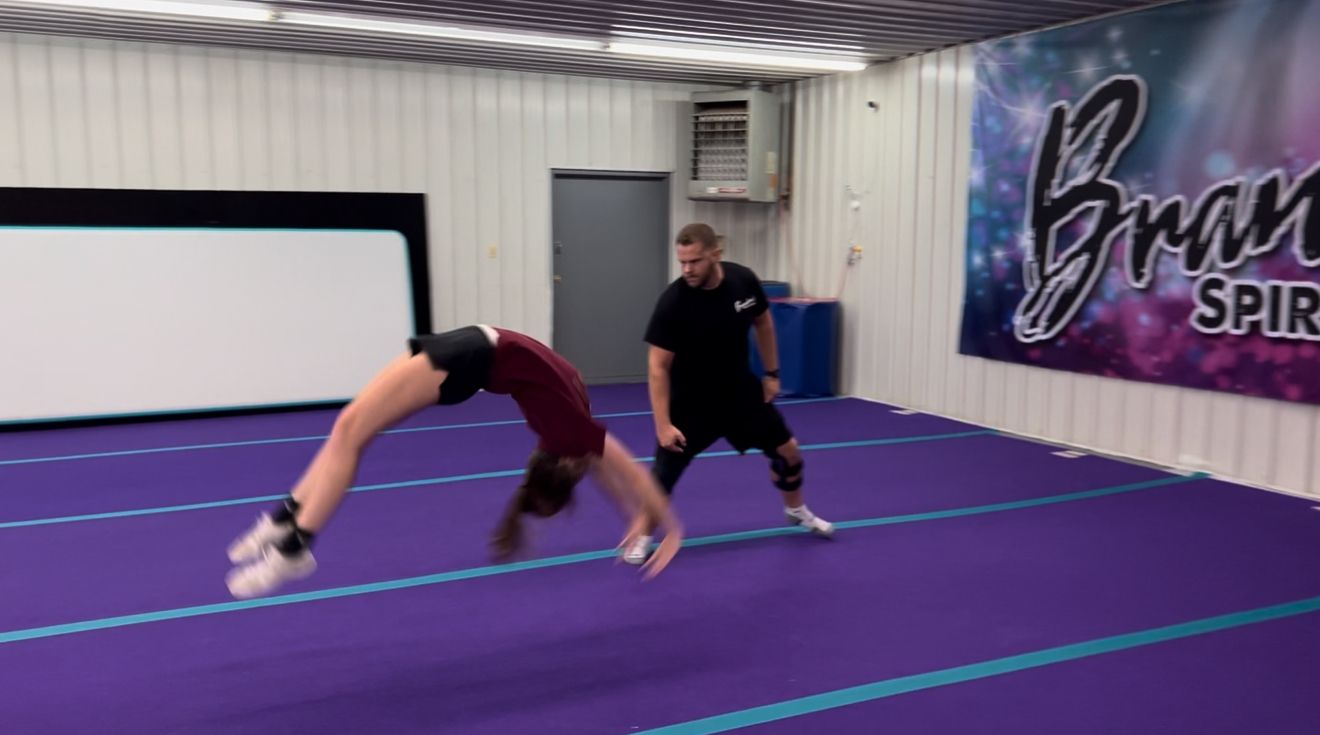 A woman in a red top performs a back handspring on a purple mat, assisted by a man in a black shirt in a gym.