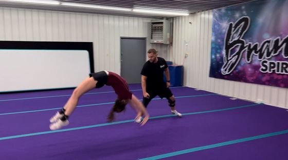 A woman in a red top performs a back handspring on a purple mat, assisted by a man in a black shirt in a gym.