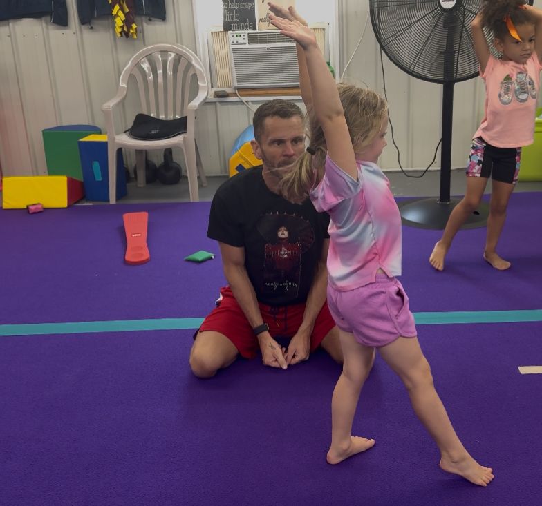 Gymnast being assisted by an instructor while practicing a move on a purple mat. Other children and a fan are in the background.