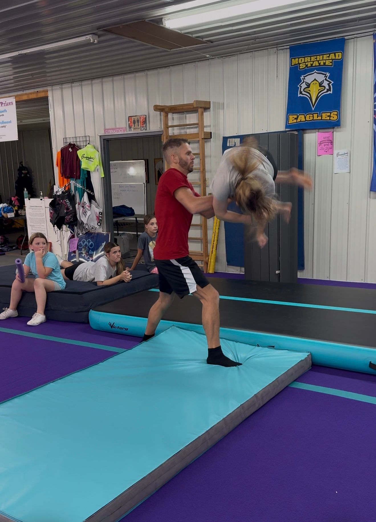 Man in red shirt assists a girl with a backflip on a mat in a gymnastics gym.