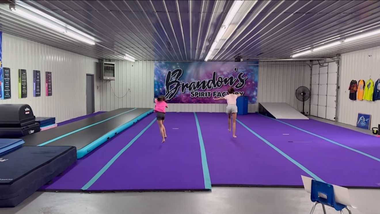 Two young girls run across a gymnastics gym with purple mats, a bouncy track, and a Brandon's All-Stars banner.