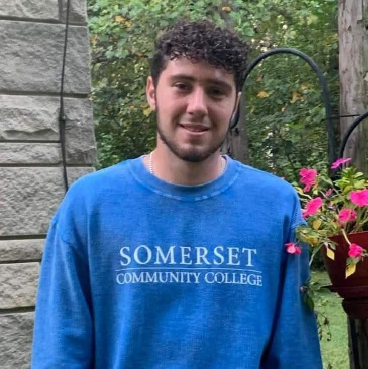 Young man with curly hair smiles, wearing a blue Somerset Community College sweatshirt. Outdoors, near a stone wall and flowers.