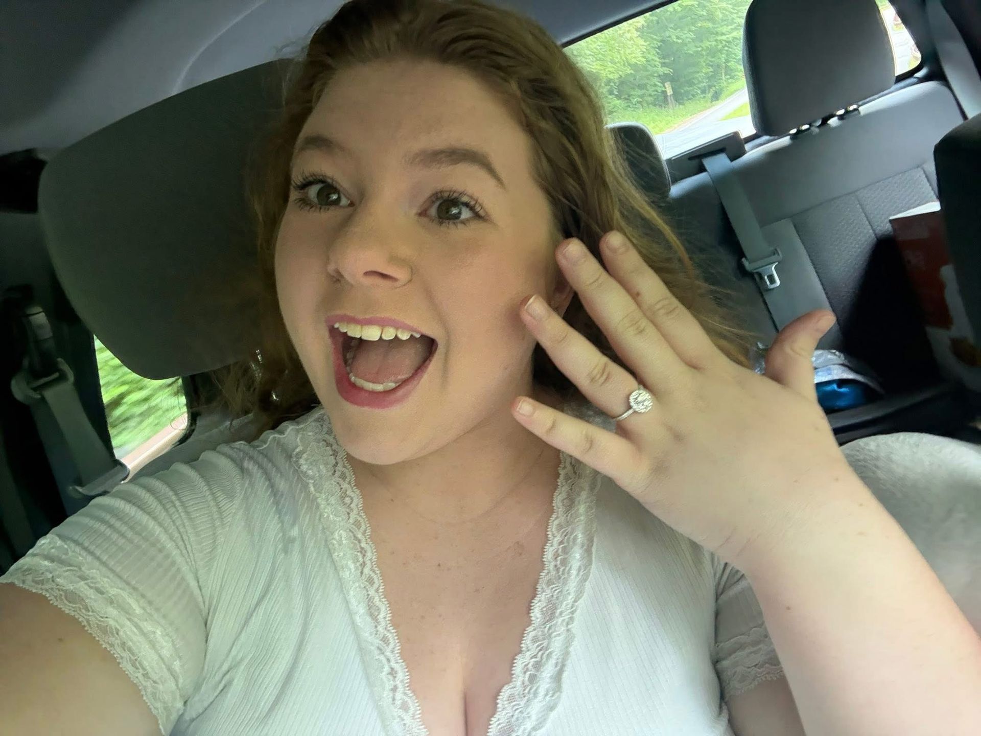 Woman in a car excitedly shows off a diamond engagement ring, smiling and looking at the camera.